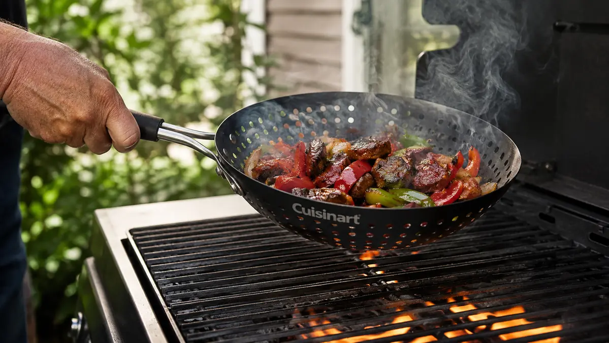 A hand shaking the Cuisinart Grill Wok to toss vegetables over a lit grill grate