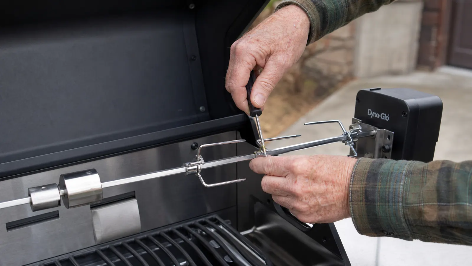 Hands locking the Dyna-Glo Universal Deluxe Rotisserie Kit's forks onto a spit rod before mounting it on a 3-burner gas grill