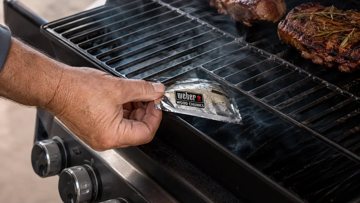 Hand placing a foil pouch of wood chunks onto a lit gas grill burner