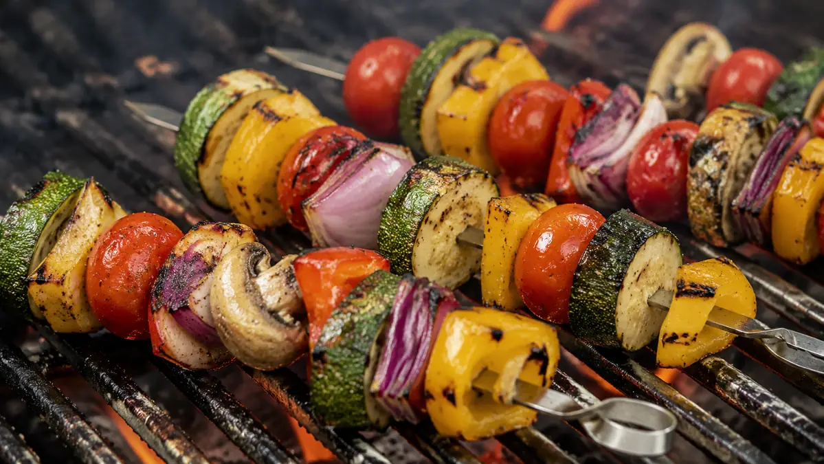 Metal skewers threaded with zucchini, peppers, and onion chunks resting across a grill grate