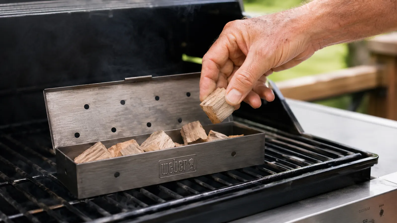 Hand placing a hickory wood chunk into a small perforated smoker box on a gas grill grate