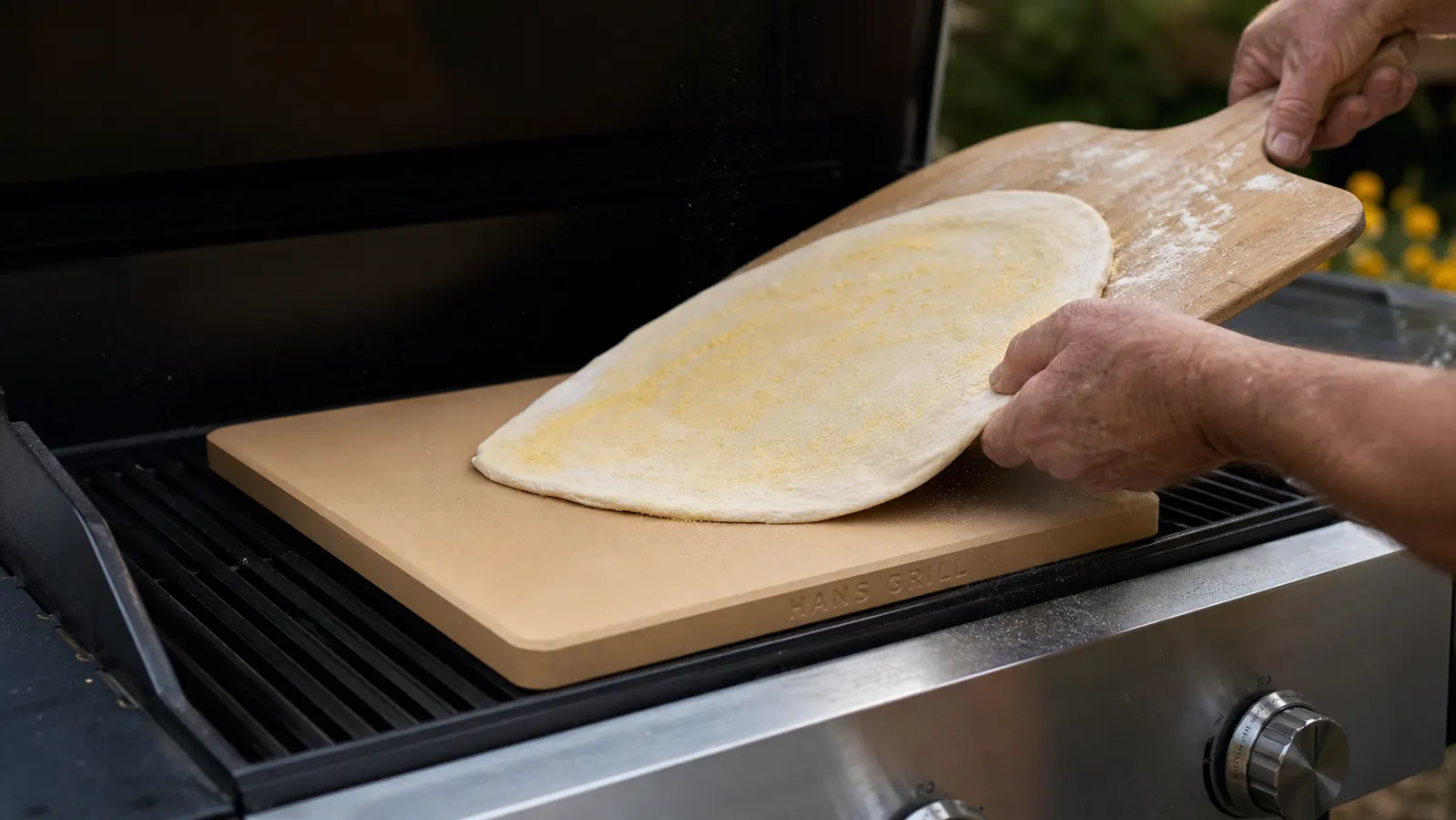Hands using a wooden pizza peel to launch a raw stretched dough onto a preheated stone on a gas grill