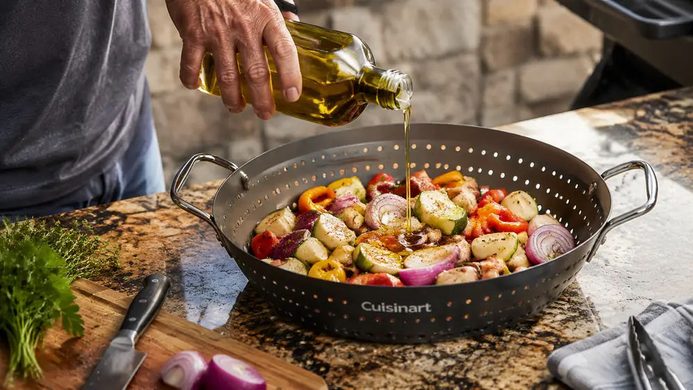 A hand pouring olive oil over diced vegetables in the Cuisinart Grill Wok before it goes on the grate