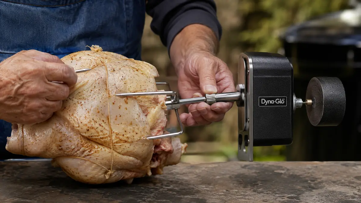 Hands sliding a rotisserie fork onto the spit rod through a trussed whole chicken