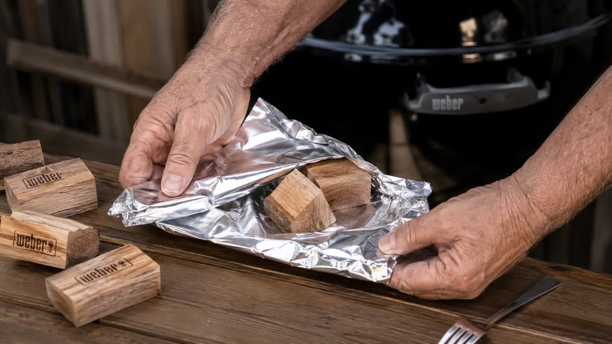 Hands wrapping hickory wood chunks in heavy-duty foil at a table beside a gas grill