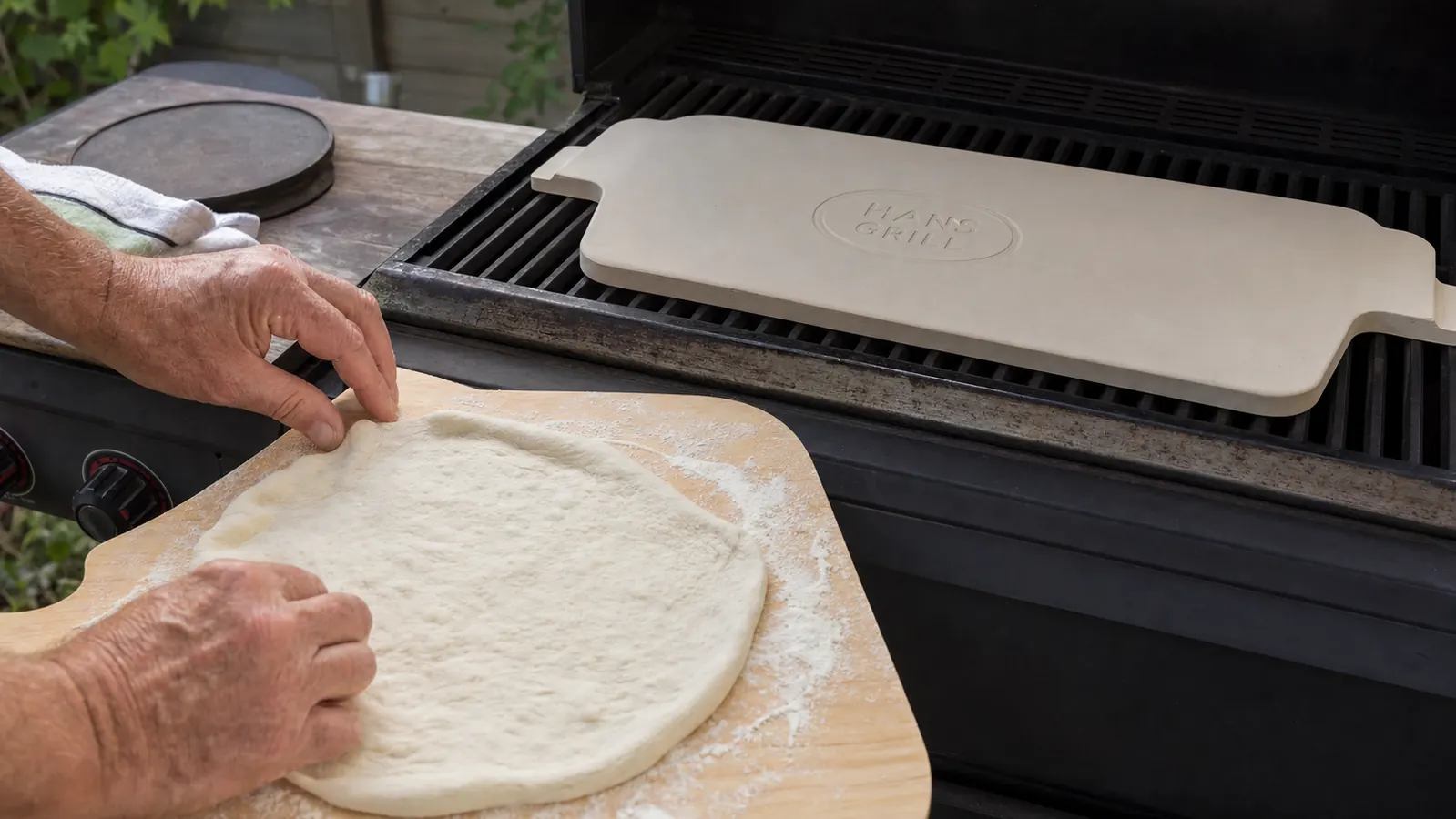 Close-up of a hand pressing raw pizza dough onto a floured wooden peel beside a preheating grill